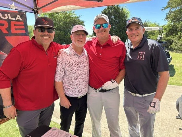 Four men posing outdoors under a canopy, with three wearing golf attire and one in casual clothes, smiling for the photo.