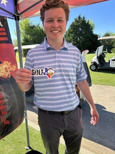 Young man smiling at the camera, wearing a striped polo shirt, holding a sticker that reads 'GOLF' with a Texas-shaped outline and Texas flag design, outdoors at a golf course or driving range with golf carts in the background.