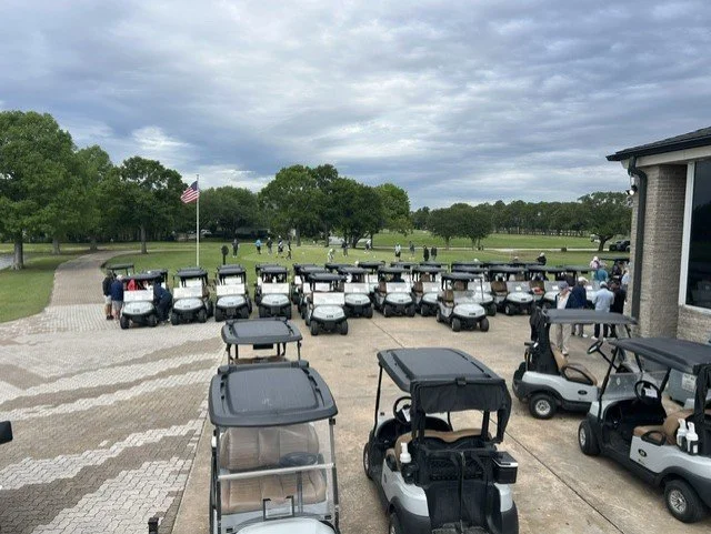 Multiple golf carts parked outside near a golf course with trees and people playing golf in the background.