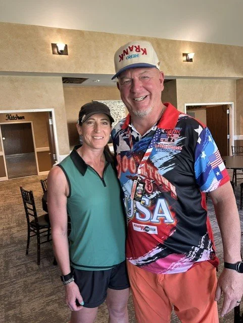 Two people, a woman and a man, standing together indoors, smiling at the camera. The woman is wearing a sleeveless green athletic top and black shorts, the man is wearing a patriotic USA-themed shirt and a white cap with red text. The background includes chairs, tables, and a sign with the word "Kitchen."