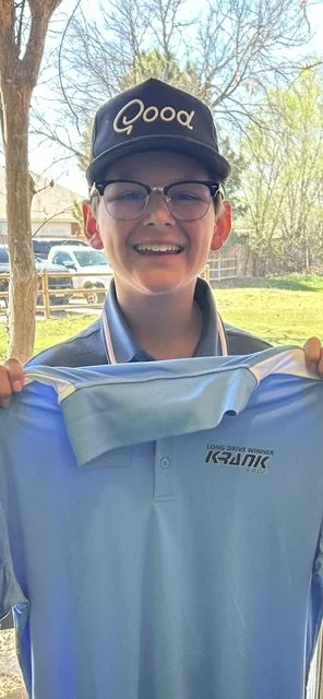 A boy with glasses wearing a black cap with the word 'Good' on it, smiling and holding a blue long-sleeve shirt outdoors with trees and cars in the background.