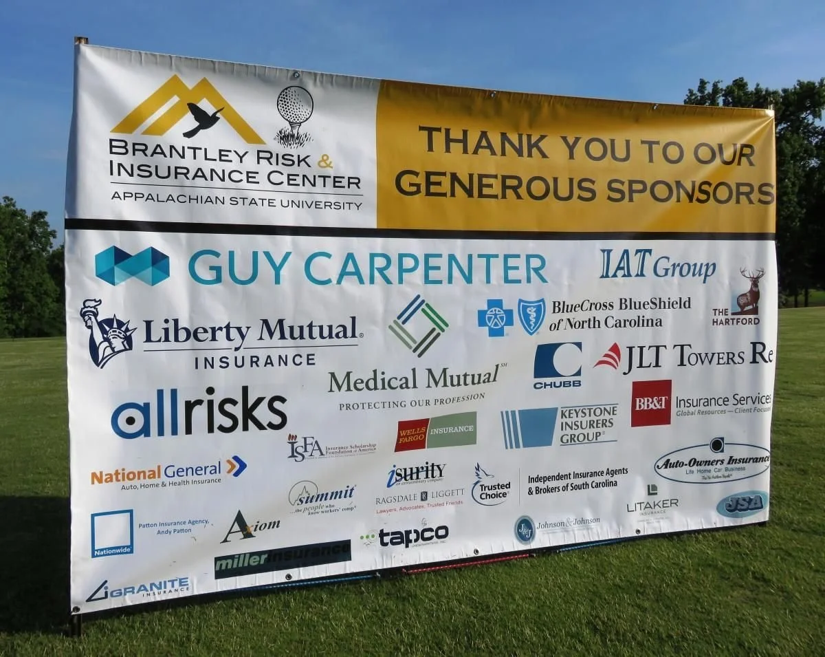 Large outdoor banner thanking sponsors of an event, featuring logos and names of multiple insurance and insurance-related companies, on a grassy field with trees and blue sky.