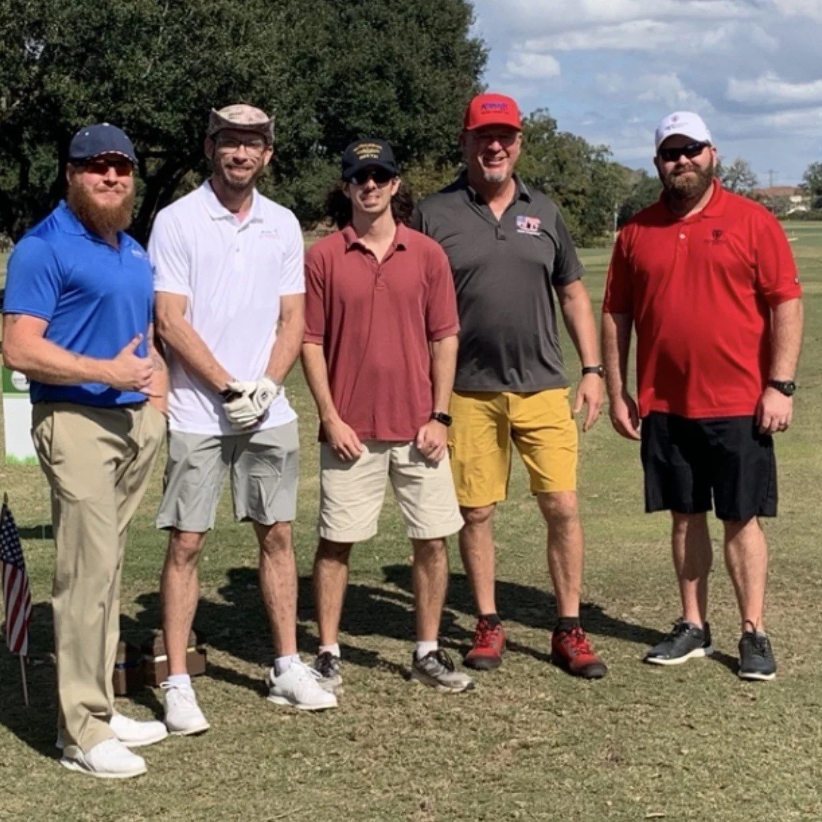 Group of five men standing on a golf course, dressed in golf attire, with trees and a partly cloudy sky in the background.