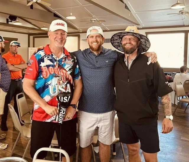 Three men smiling and posing together in a room, with other people in the background. The man on the left is wearing a colorful patriotic shirt and a white cap, holding a golf club. The man in the middle is wearing a blue polo shirt and a white cap. The man on the right is wearing a black jacket, a wide-brimmed hat, and shorts, with tattoos on his arms.