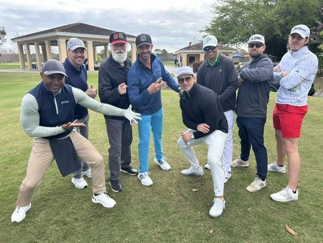 Group of nine people on a golf course posing together, some making gestures, with a pavilion and trees in the background.