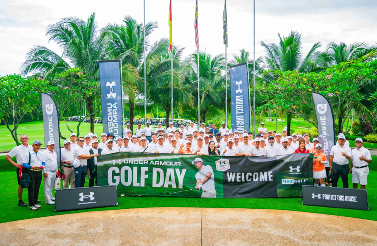 Group of people participating in an Under Armour golf event on a golf course, holding a large banner that reads "GOLF DAY" and "WELCOME" with lush green trees and flags in the background.