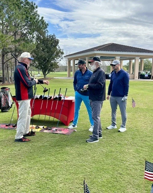 Four men in golf attire standing on a golf course, one person showing golf clubs at a display table.