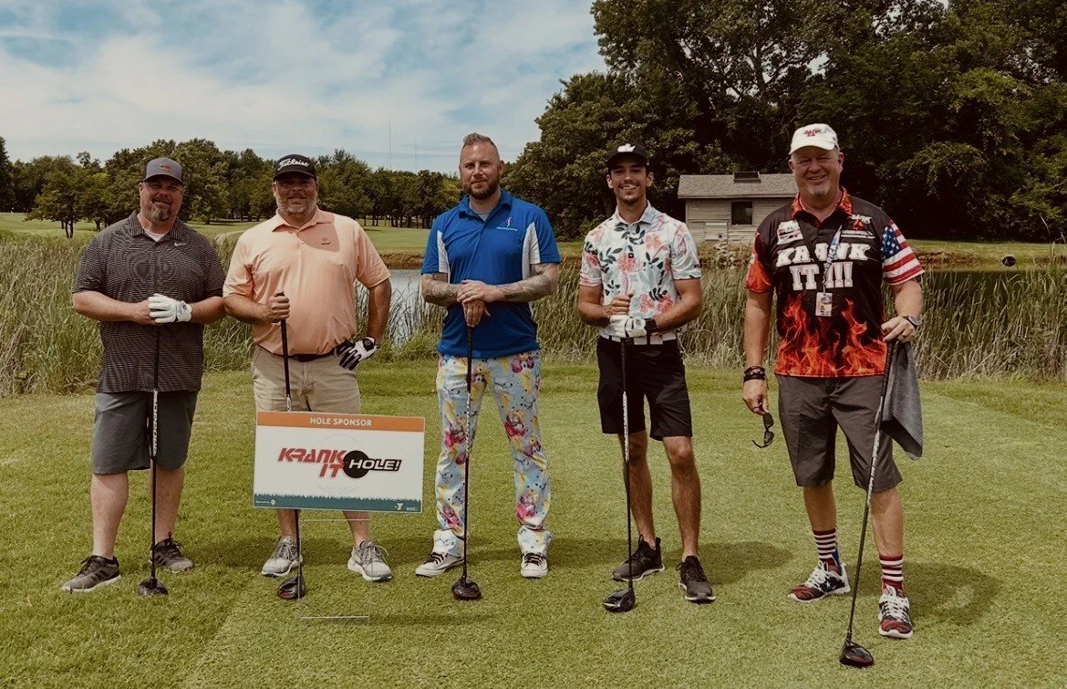 Five men standing on a golf course holding golf clubs, with a sign that reads 'HOLE SPONSOR KRANK IT OUT HOLE.' They are dressed in golf attire, smiling, with trees, grass, and a pond in the background.
