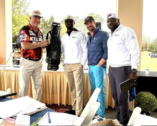 Four men standing together outdoors, two holding items, smiling at the camera, at a golf event with a table in front and trees in the background.