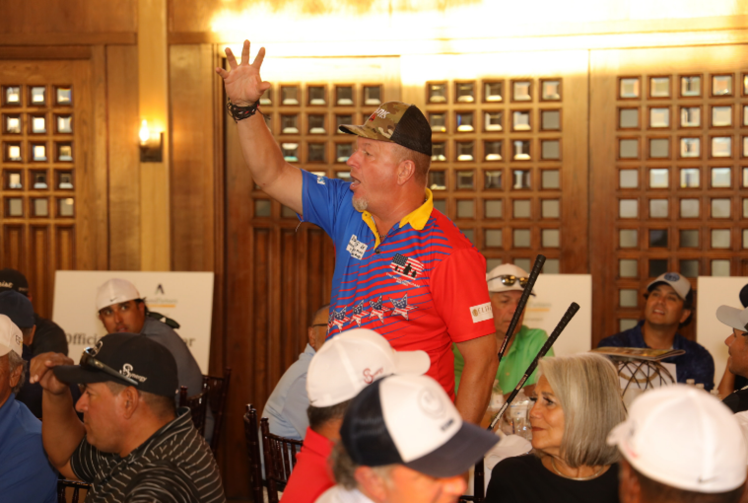 A man in a colorful polo shirt with a camouflage hat, standing and raising his hand in a crowded indoor setting with wooden decor, surrounded by seated people wearing caps.