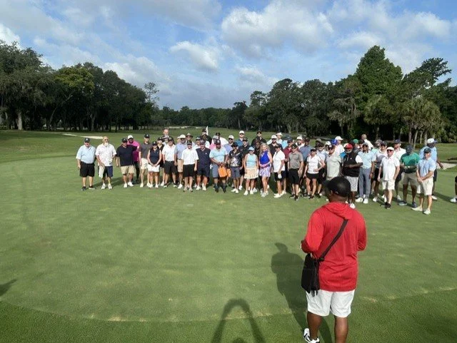 Group of people gathered on a golf course, with a person in a red shirt and white shorts standing in the foreground.