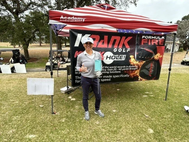A woman standing on a golf course holding a trophy and a box in front of a promotional tent for a golf ball brand called 'KRANK'. The tent has a banner with the brand and a golf-related graphic.