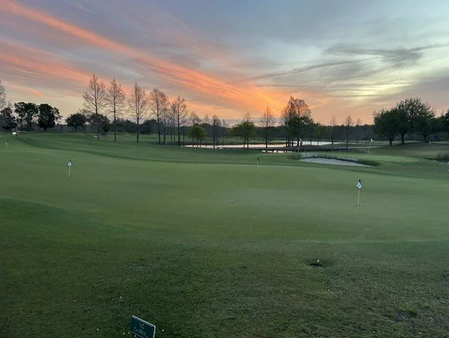 A golf course at sunset with a water hazard, green grass, and trees in the background.