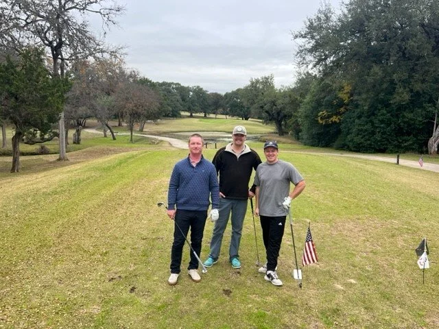 Three men standing on a golf course with golf clubs, surrounded by trees, during a cloudy day.