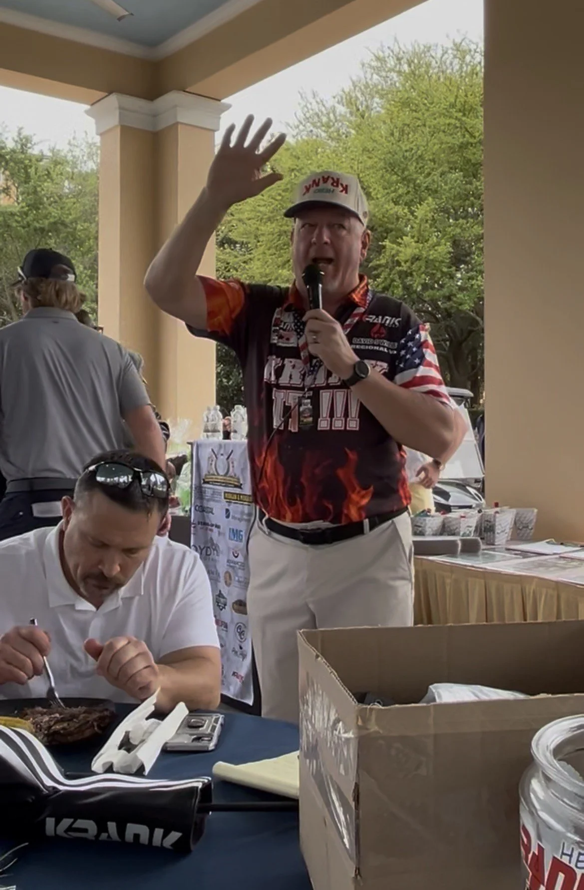 A man wearing a motorcycle racing jersey and a cap with 'KRAUK' raises his hand while speaking into a microphone at an outdoor event. There are people around, some sitting at a table with food and papers, and trees in the background.