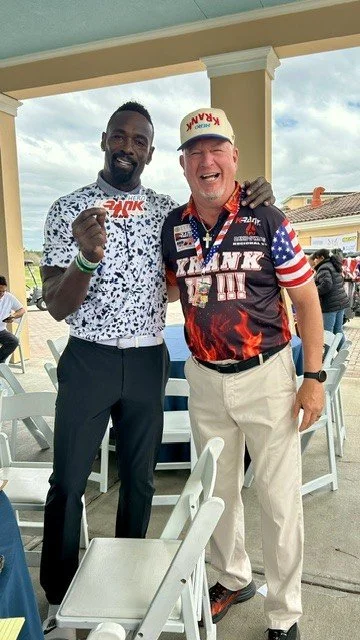 Two men standing outdoors, smiling and posing for the camera. One is holding a white and red checkered plate or object, the other has his arm around the first man's shoulder. They are dressed casually and seem to be at a social event or gathering.