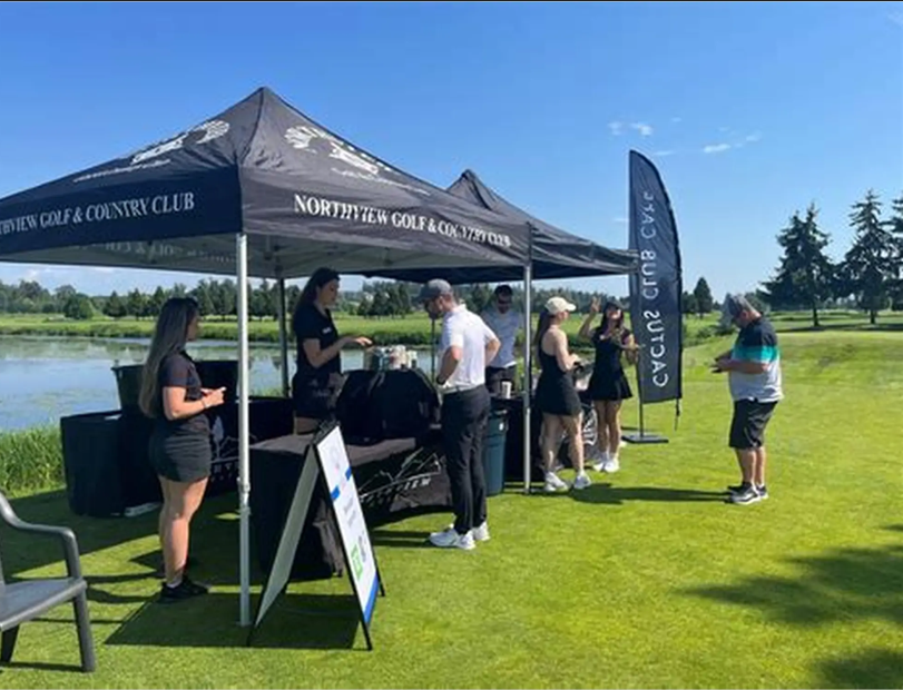 People gathering at a canopy tent on a golf course, with some ordering drinks or snacks, under a clear blue sky near a pond.