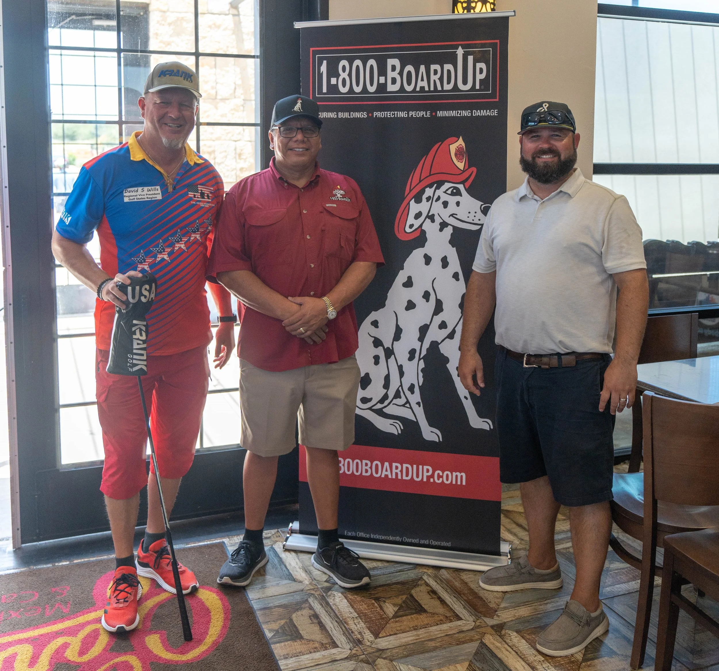 Three men standing in front of a black and red banner for Board Up, a business specializing in construction site protection. The man on the left is wearing a colorful golf shirt, shorts, and orange and black sports shoes. The man in the middle is wearing a red shirt, dress shorts, glasses, a cap, and sneakers. The man on the right is wearing a white polo shirt, dark shorts, sunglasses on his cap, and beige shoes. They are indoors near a large window and a colorful carpet.