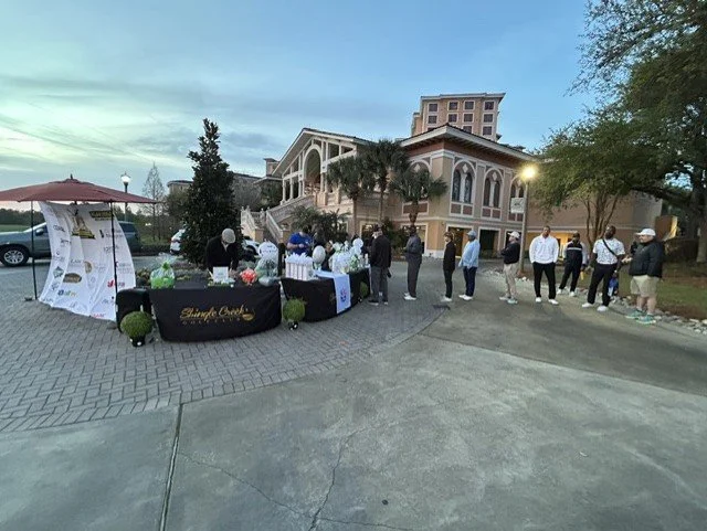 People waiting in line outside a building for an event or service during the evening, with tables and promotional materials set up nearby.
