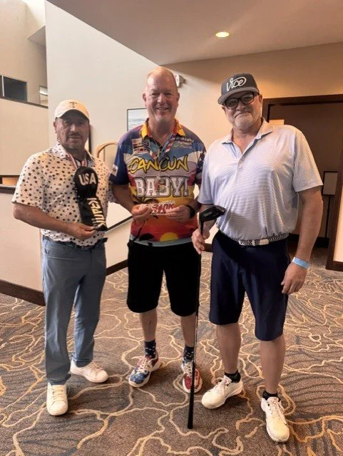Three men standing indoors, smiling, one holding a golf club, wearing casual golf attire including hats and sunglasses.