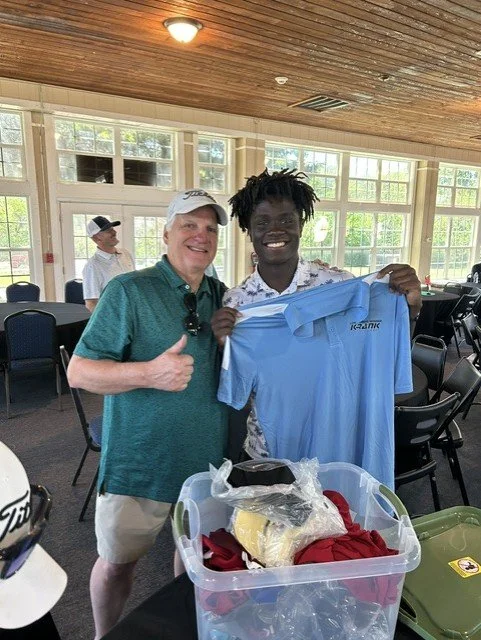 Two men smiling and holding a blue polo shirt in a room with large windows. One man is giving a thumbs-up and the other is showing the shirt. There are tables and chairs in the background and a plastic storage bin with various items on the table.