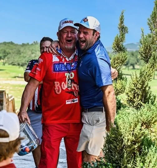 Two men hugging and laughing outdoors in a golf course setting, surrounded by greenery and trees, with other people in the background.