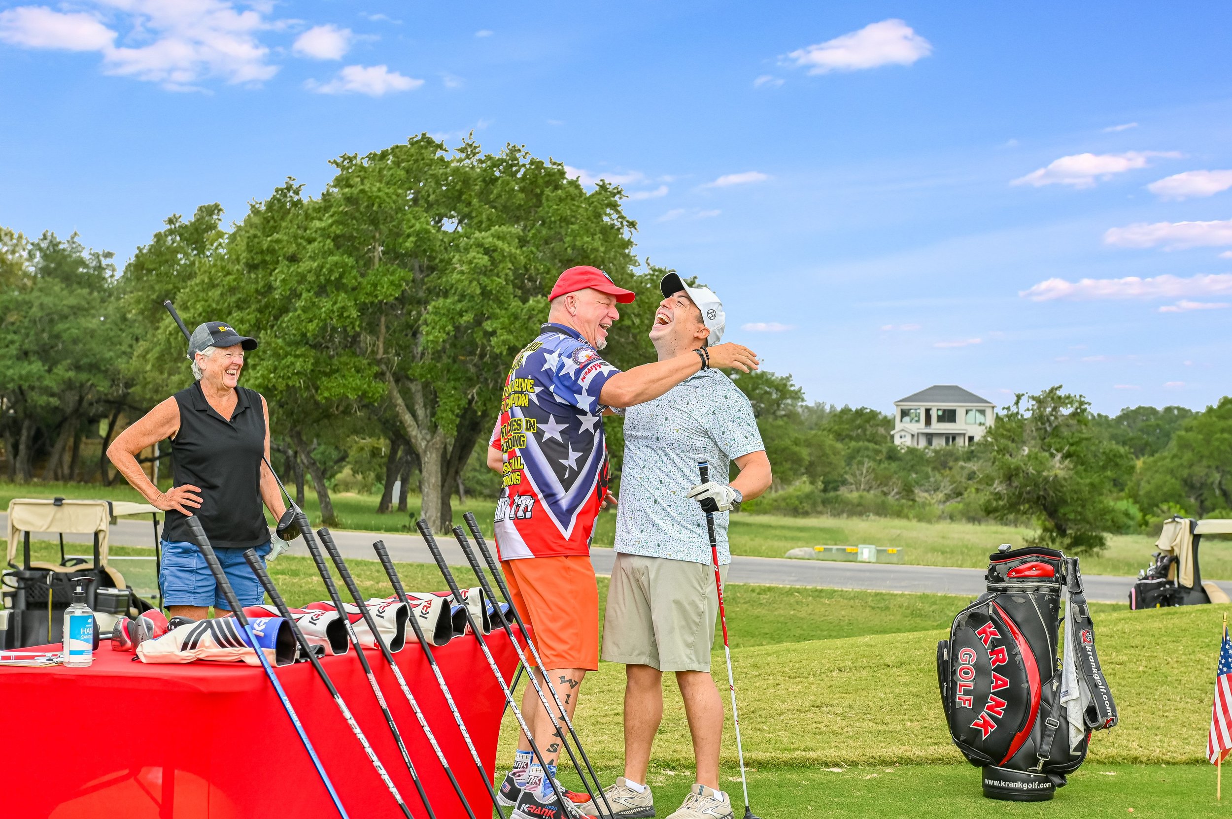 Two men are smiling and hugging on a golf course, surrounded by golf equipment and bags. A woman stands nearby, smiling. The scene takes place outdoors on a sunny day with trees and a house in the background.