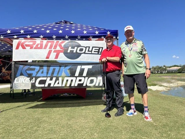 Two men standing on a golf course, with an American flag-patterned canopy behind them. One man in a red polo shirt and black pants is holding a golf club, and the other in a green shirt and dark shorts is standing next to him. There are banners reading "KRANK IT" and "CHAMPION" in the background.