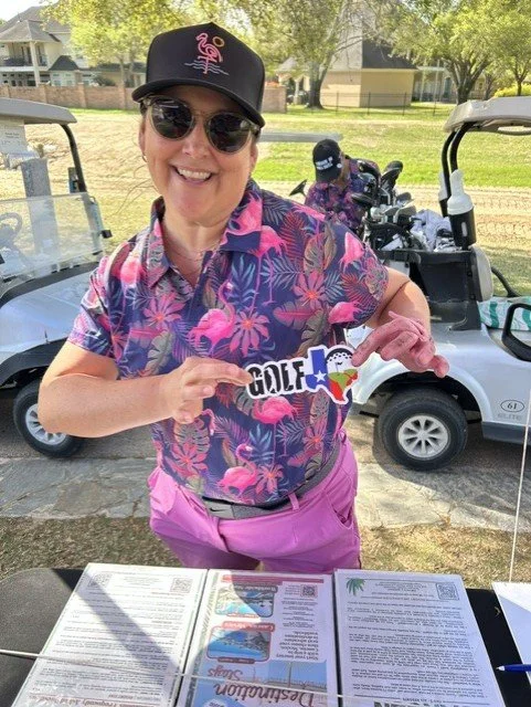A cheerful woman wearing sunglasses, a black golf cap, and a colorful floral shirt holding a decorative golf-themed sign, standing outdoors near golf carts.