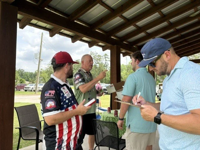 Four men gathered outdoors under a wooden pavilion, one of whom is speaking while the others listen and take notes.