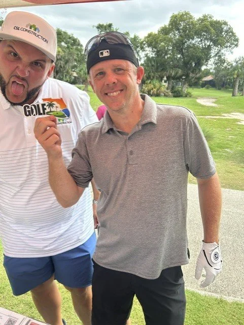 Two men on a golf course holding a sign that says 'GOLF'. The man on the left is wearing a white cap and a white shirt, sticking his tongue out. The man on the right is wearing a black cap, a gray polo shirt, and golf gloves, smiling at the camera.