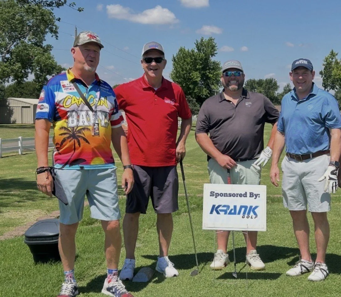 Four men standing on a golf course, smiling, holding golf clubs, wearing golf attire, with a sign that says 'Sponsored By: Krank Golf'.