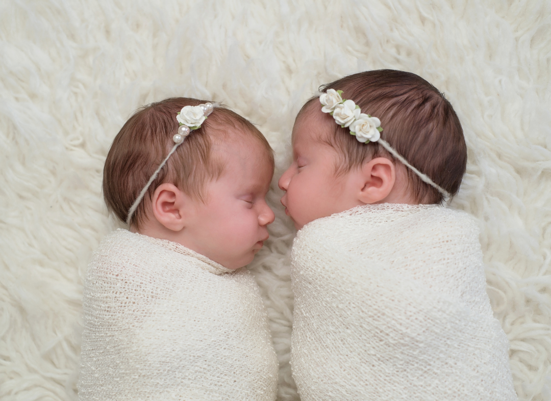 Two newborn babies swaddled in white blanket, lying on a soft furry surface, wearing floral headbands, facing each other with eyes closed.