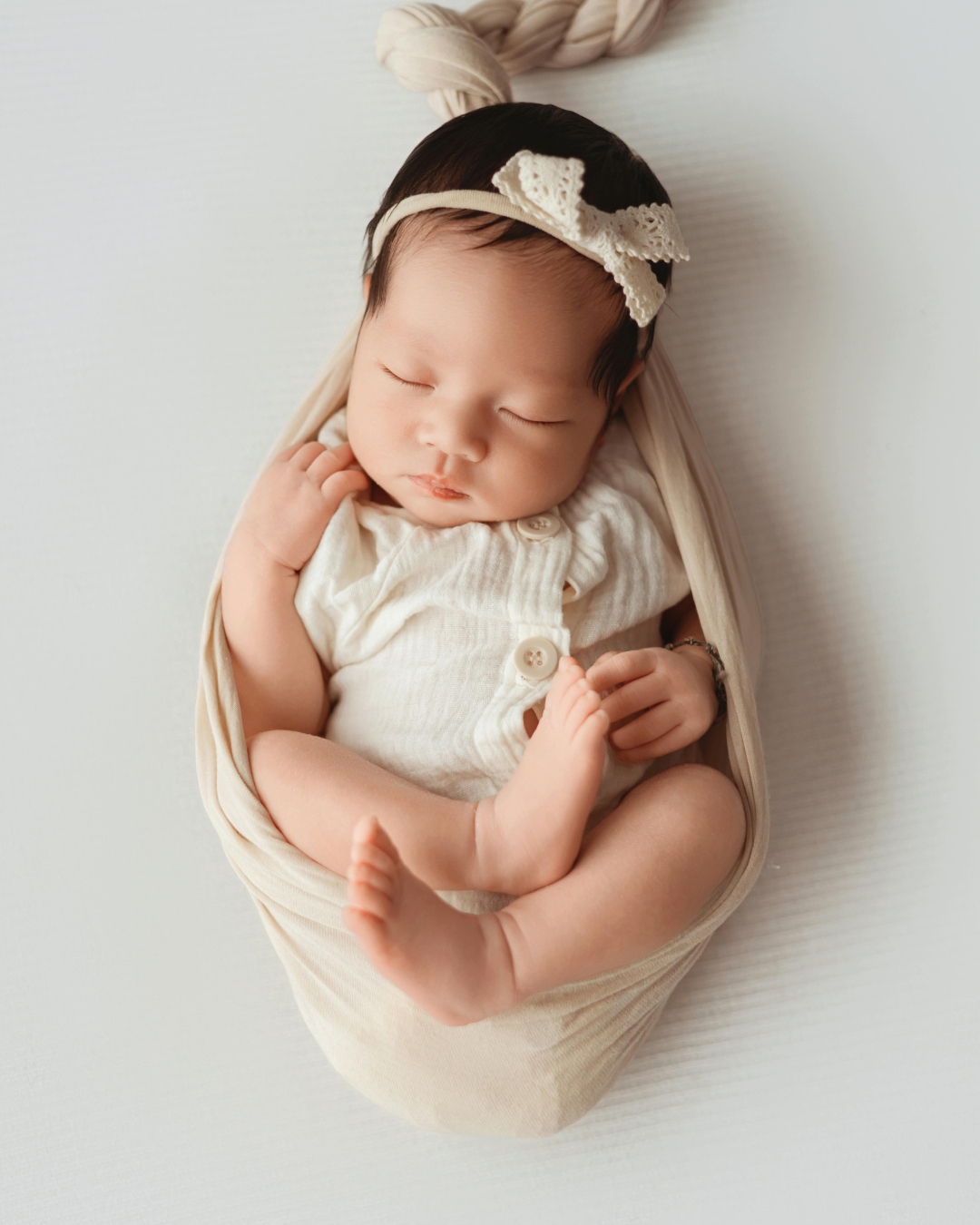 Close-up of a peacefully sleeping baby with a headband and lace bow, wrapped in a cream-colored swaddle on a white surface.