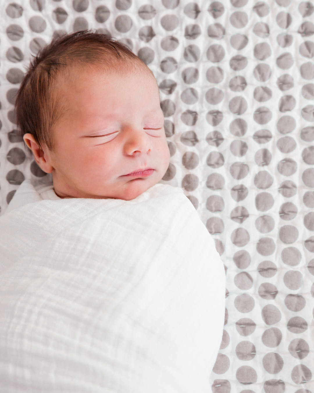 A sleeping newborn baby wrapped in a white blanket on a polka dot sheet.