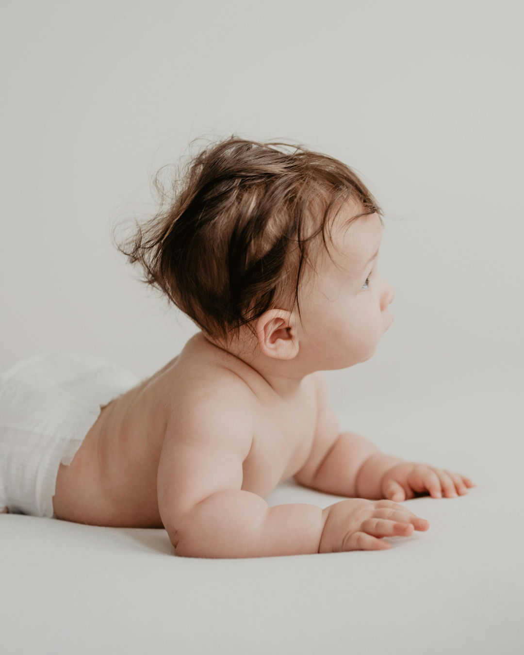 Side view of a baby lying on their stomach with brown hair, looking to the right, on a plain white surface against a plain wall.