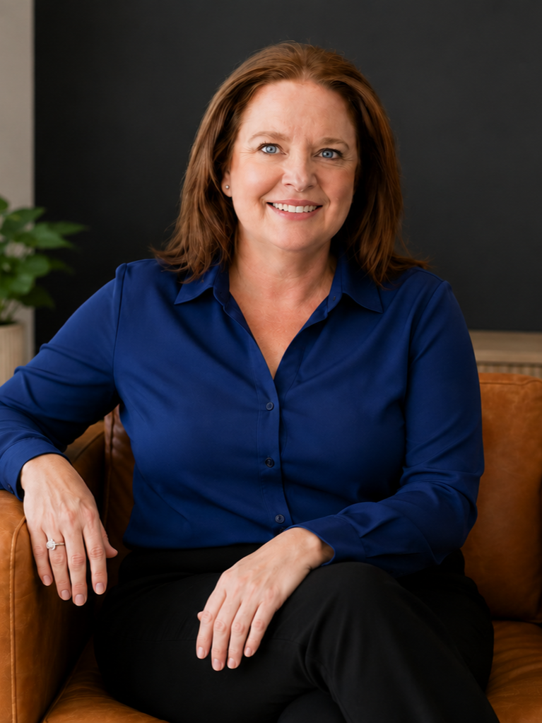 A woman with red hair and blue eyes sitting on a brown leather couch, smiling, wearing a blue button-up shirt and black pants, with a plant and a dark wall in the background.