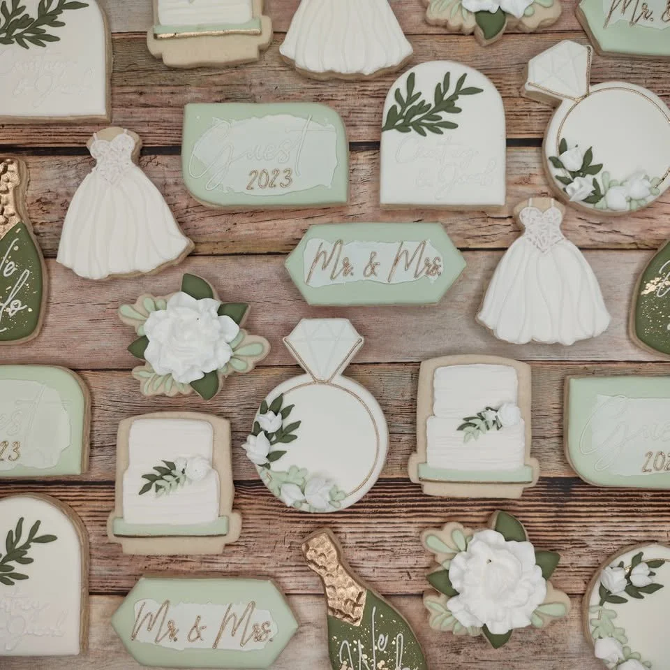 Decorative wedding cookies with floral and wedding dress designs, labeled 'Mr. & Mrs.', 'Guest 2023', and other wedding-themed decorations on a wooden surface.