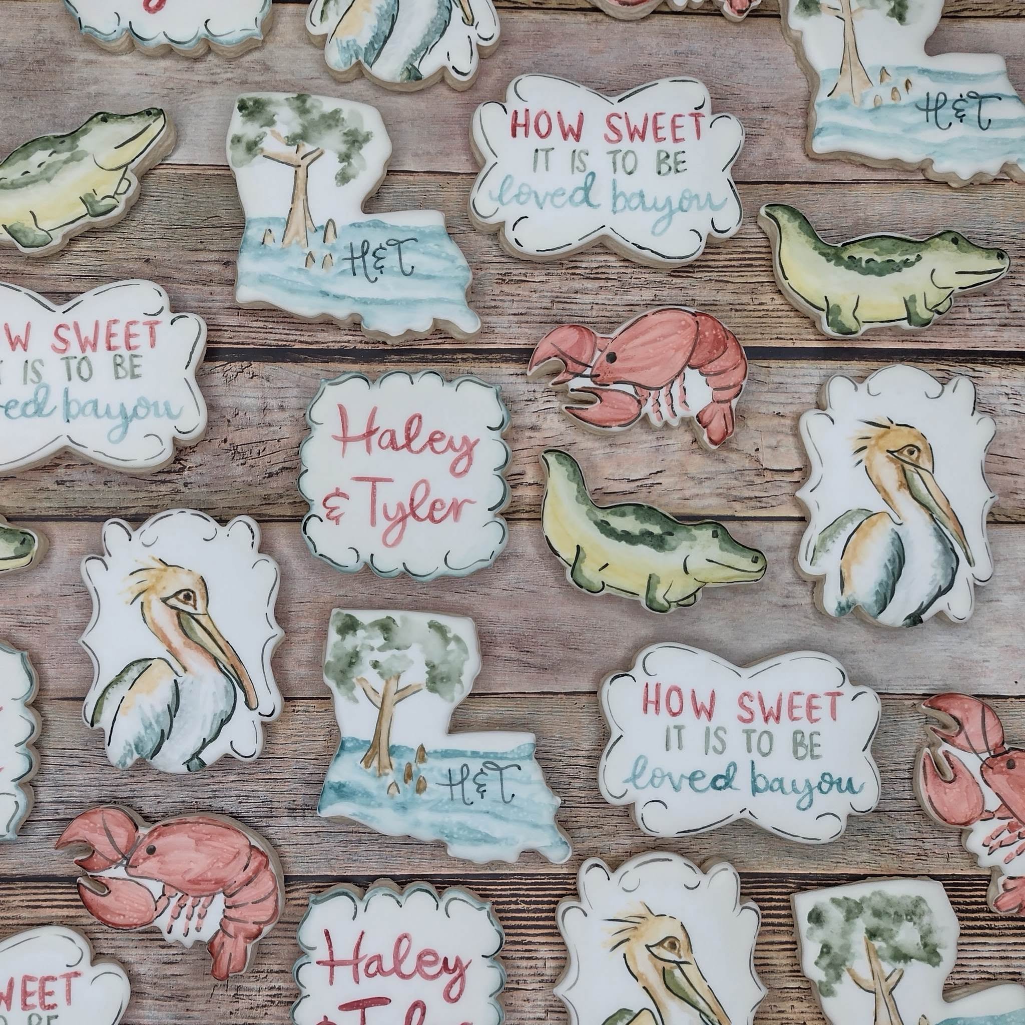 Collection of decorated cookies with various themes including cypress trees, animals like crocodiles, crawfish, herons, and messages such as 'How sweet it is to be loved by you' on a wooden surface.