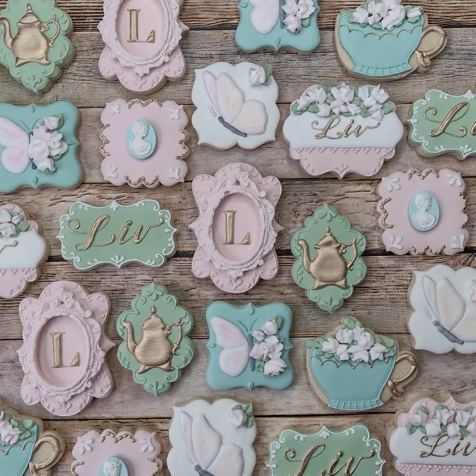 Tea party decorated cookies with pastel colors, floral and tea cup motifs, butterfly, and letter L, placed on a wooden surface.