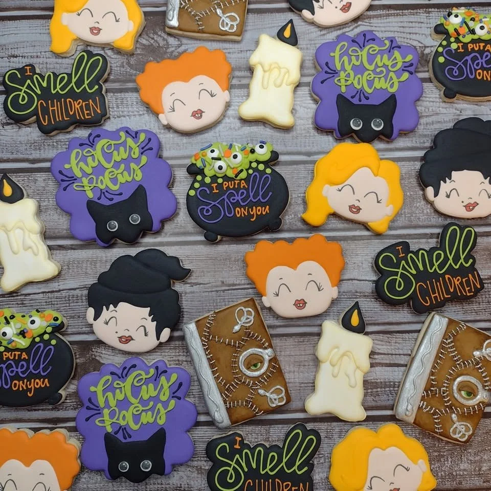 Hocus Pocus-themed decorated halloween cookies including witches, candles, black cats, spell book, and phrase cookies with sayings like "I smell children" and "I put a spell on you" on a wooden surface.