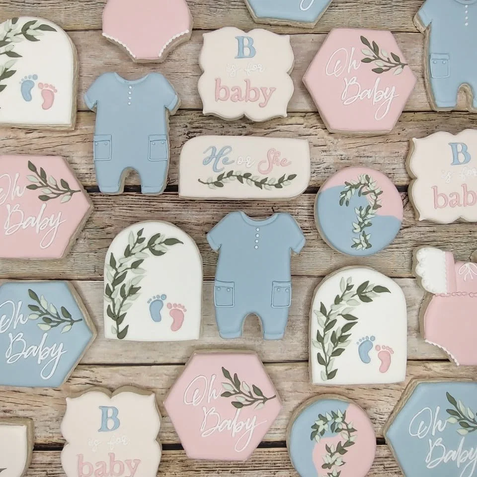 Decorative cookies with baby themes in pastel colors, including onesies, baby footprints, 'Oh Baby' and 'He or She' text, and floral and leafy designs on a wooden surface.