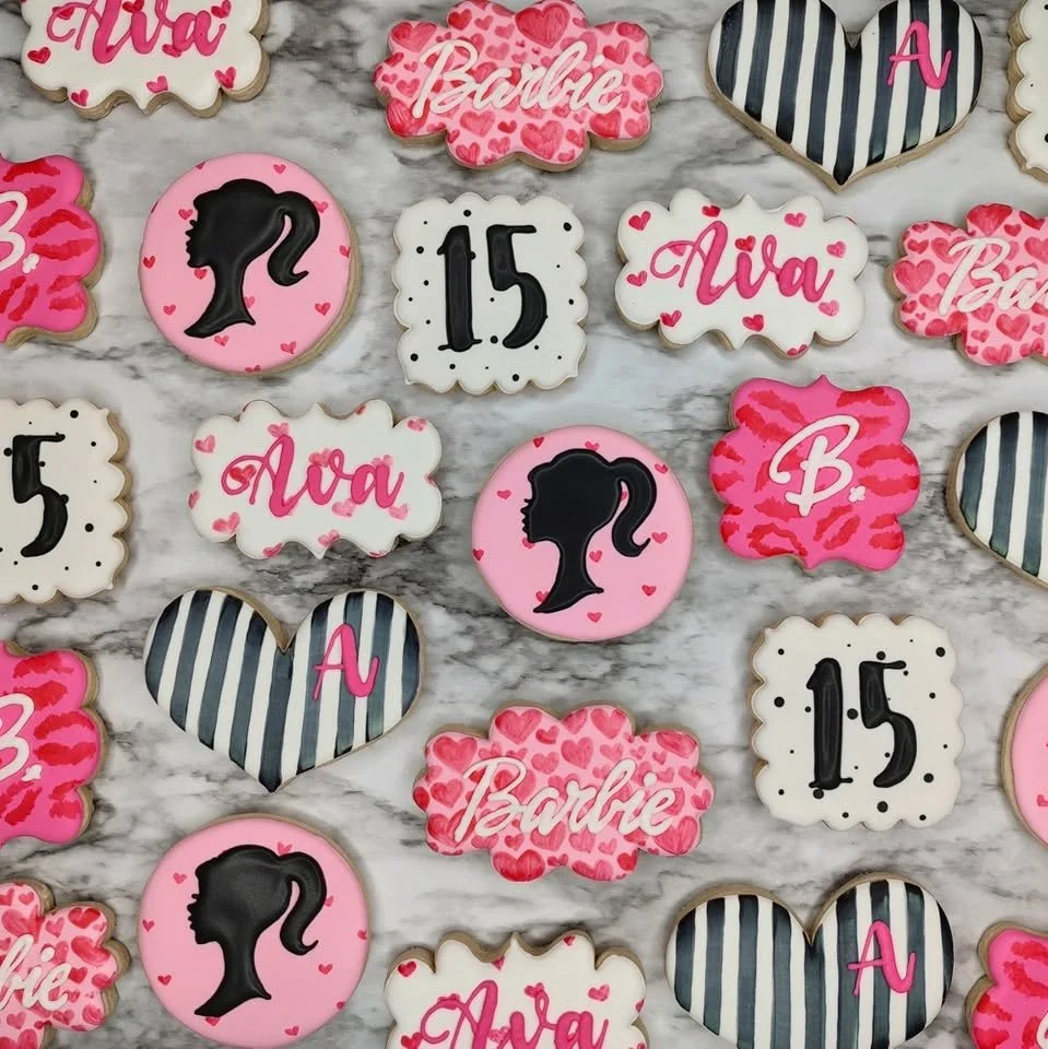 Barbie themed decorated cookies with various shapes and designs, including hearts, featuring text such as 'Ava,' 'Barbie,' and the number '15,' with pink, black, and white colors, placed on a marble surface.