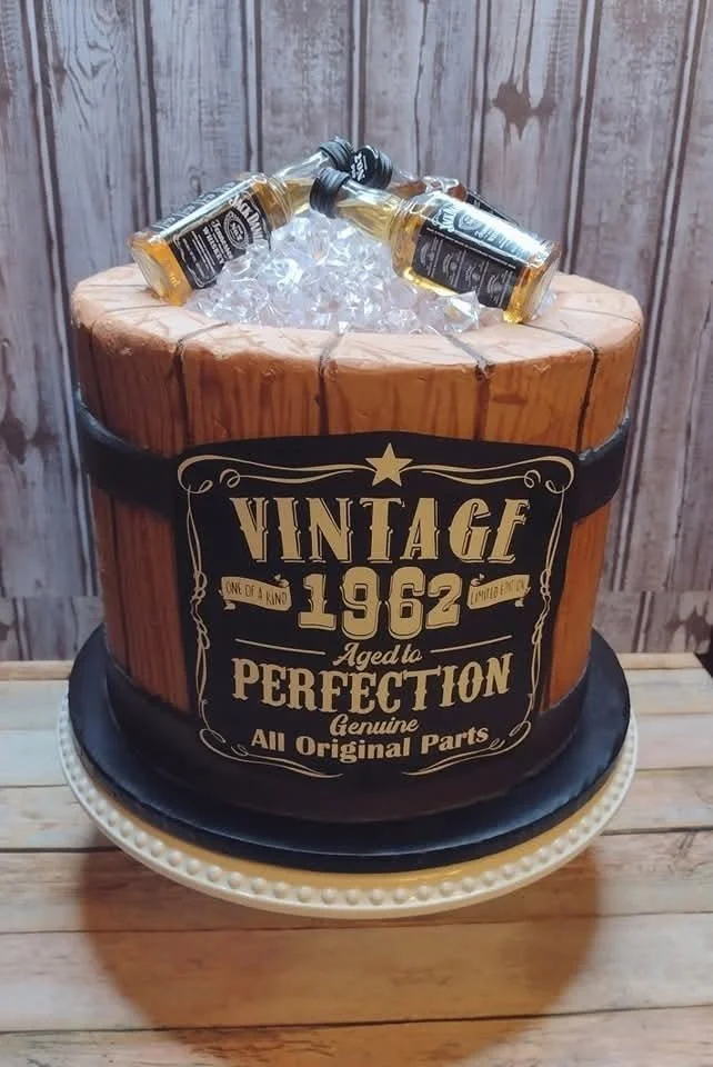 A birthday cake designed to look like a vintage wooden barrel with a black band around it, featuring the words 'VINTAGE 1962 Aged to PERFECTION' and other decorative text. The cake has small bottles of Jack Daniel's whiskey on top.
