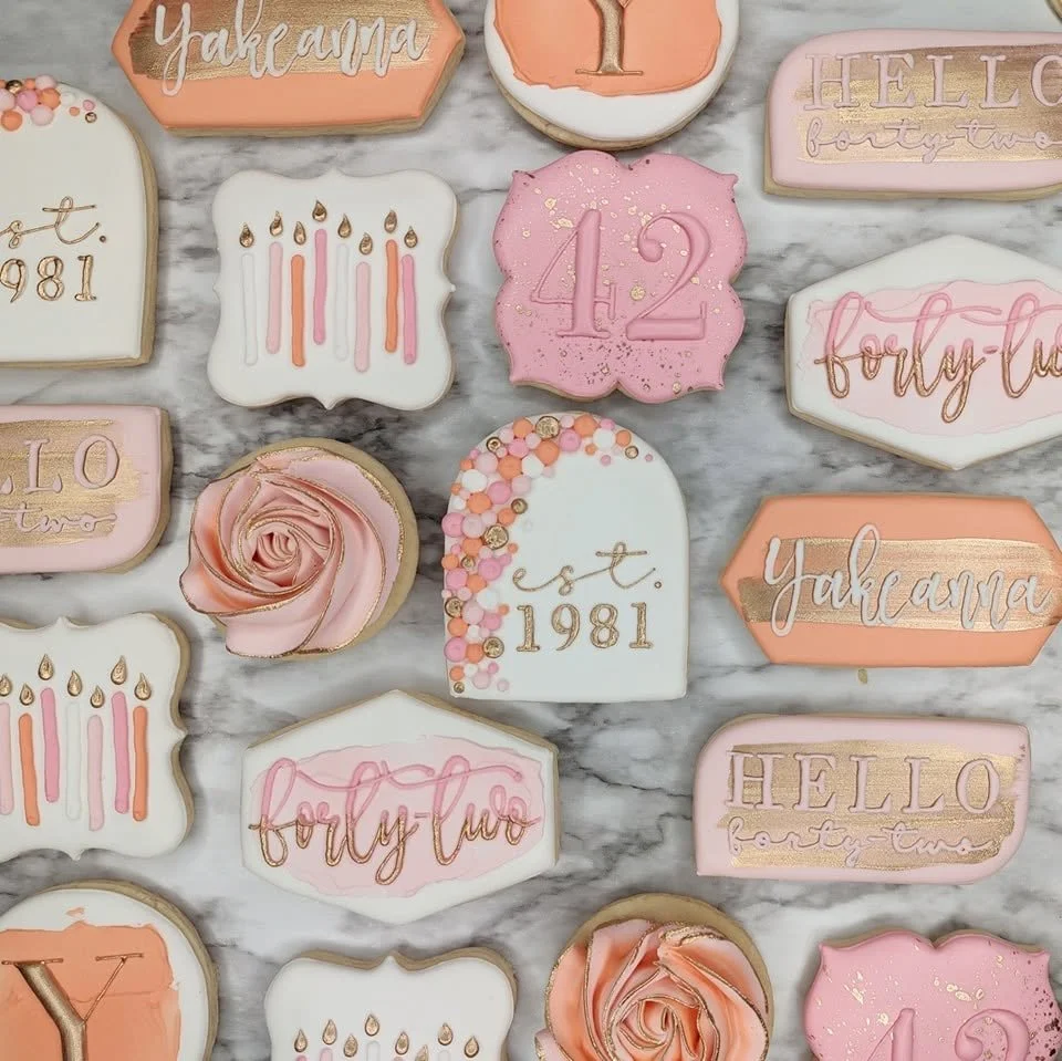 Decorated cookies celebrating a 42nd birthday with pink, white, and peach tones, featuring number 42, candles, roses, and phrases like "Hello forty-two" and "forty-two" on a marble surface.