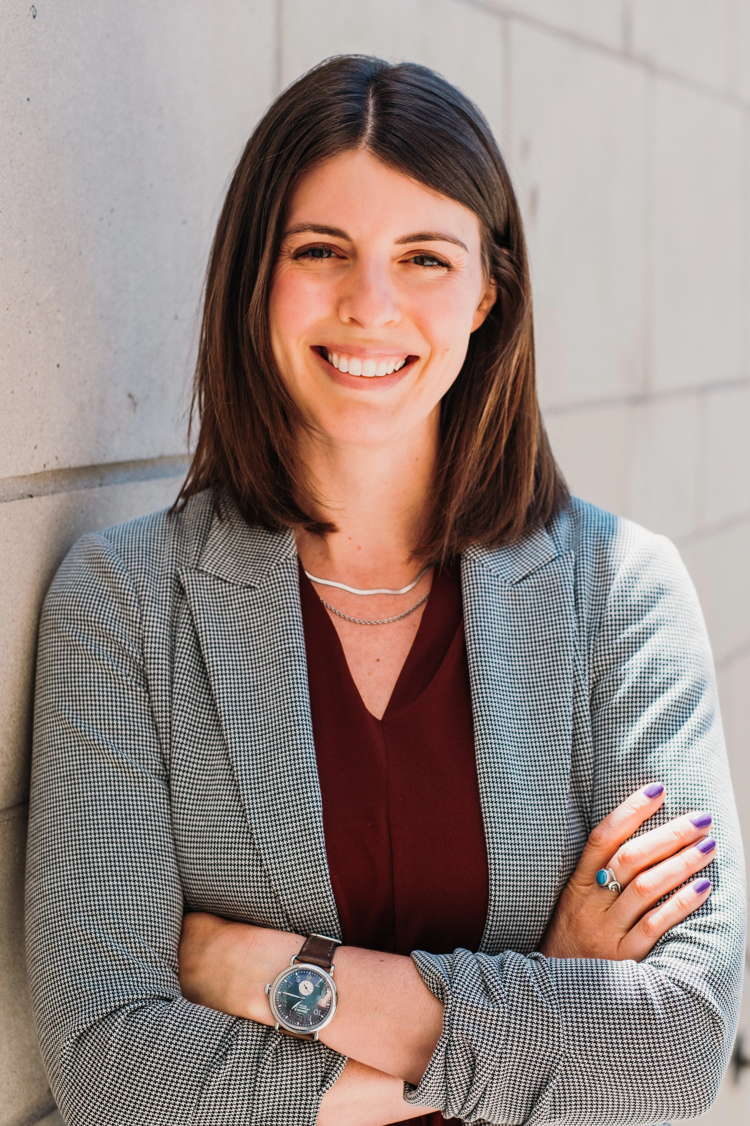 A woman with shoulder-length brown hair, crossed arms, wearing a watch, rings, a plaid blazer, and a maroon top, leaning against a concrete wall and smiling.