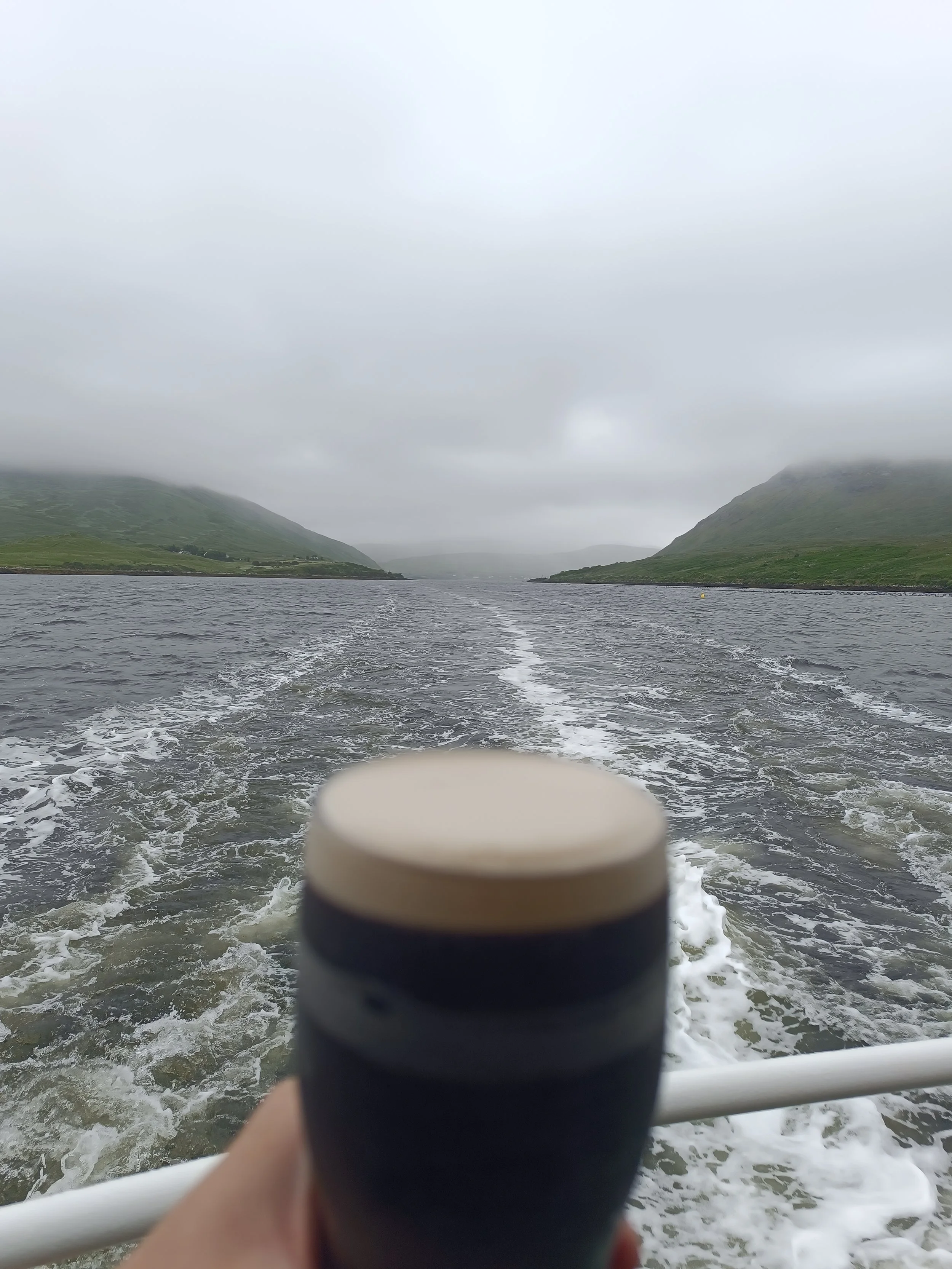 Photo taken from the back of a boat or ferry traveling on a body of water. The water is choppy, with small waves. In the foreground, part of the boat or ferry's railing is visible, with a finger or hand holding onto it. The background features a landscape of green, grassy hills or mountains shrouded in mist or fog, with a cloudy, overcast sky.