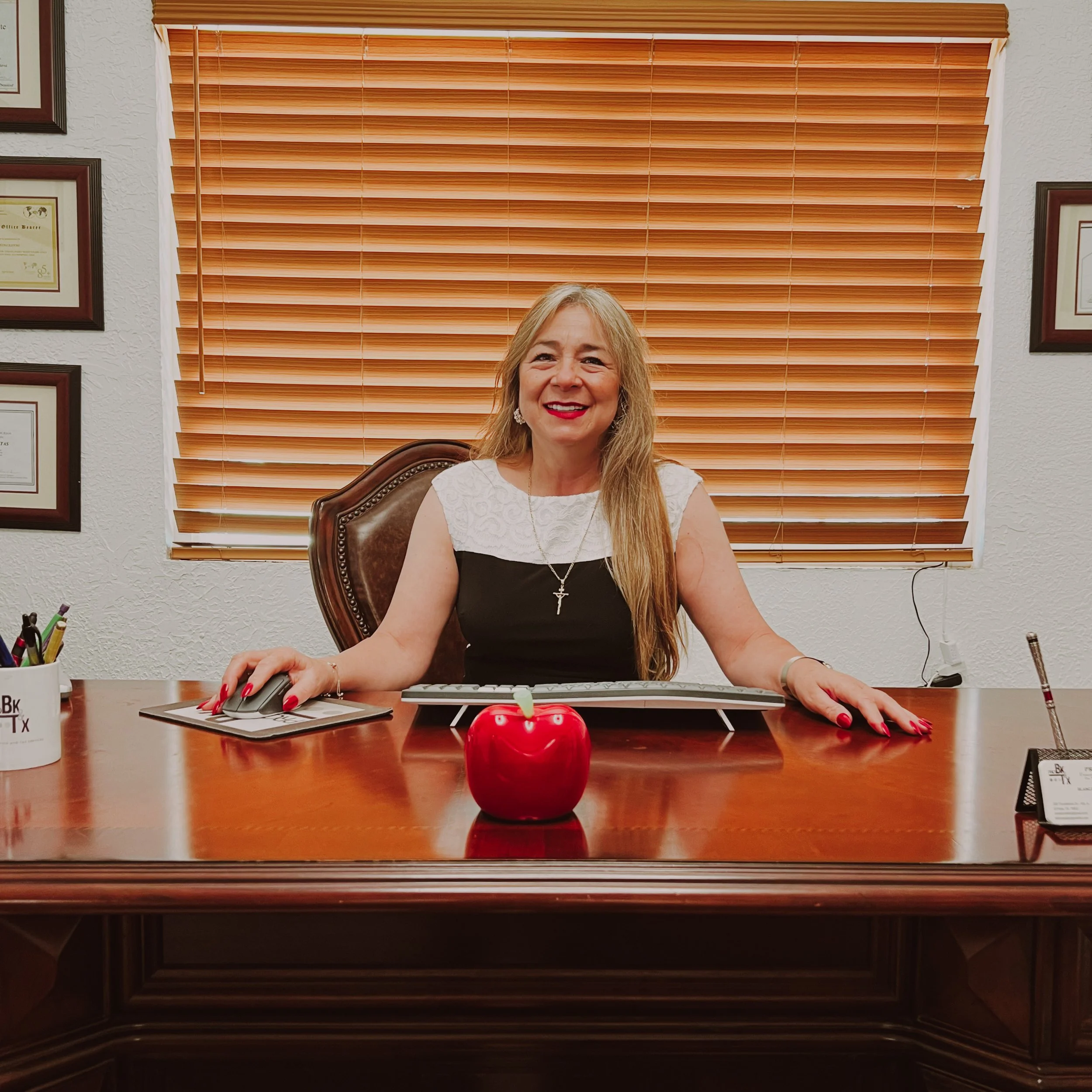 PROBKTX Director with long blonde hair sitting at a desk with a keyboard, mouse, red apple, and pen holder. Behind her is a large window with closed wooden blinds. The wall is decorated with framed certificates or diplomas.