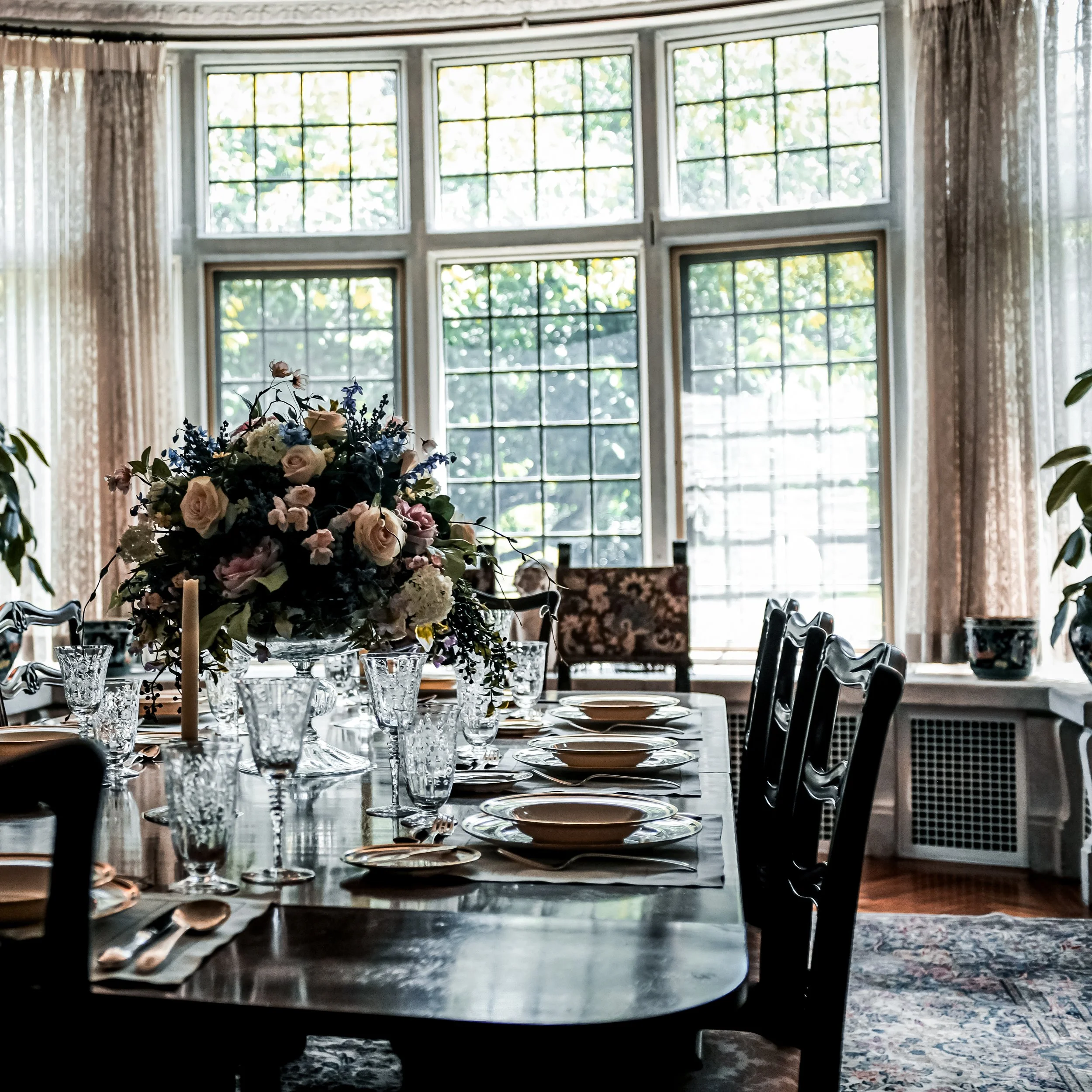 A dining room with a large window, decorated with flowers on the table, set with glasses, plates, and silverware.