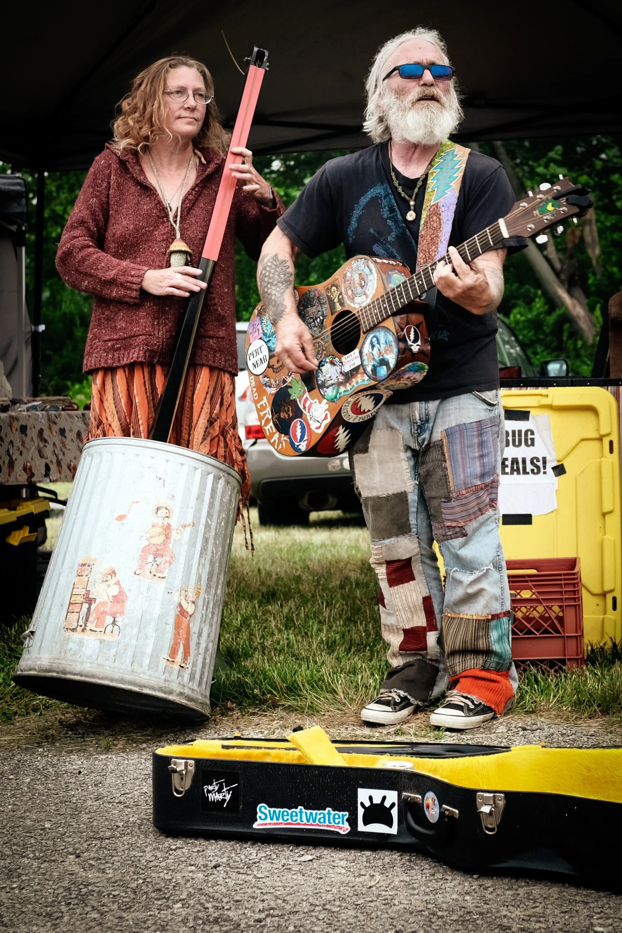 A man with a white beard and sunglasses playing an acoustic guitar decorated with stickers, standing next to a woman holding a large, vintage metal bucket with a painted design, both on a grassy outdoor stage with green trees and equipment in the bac